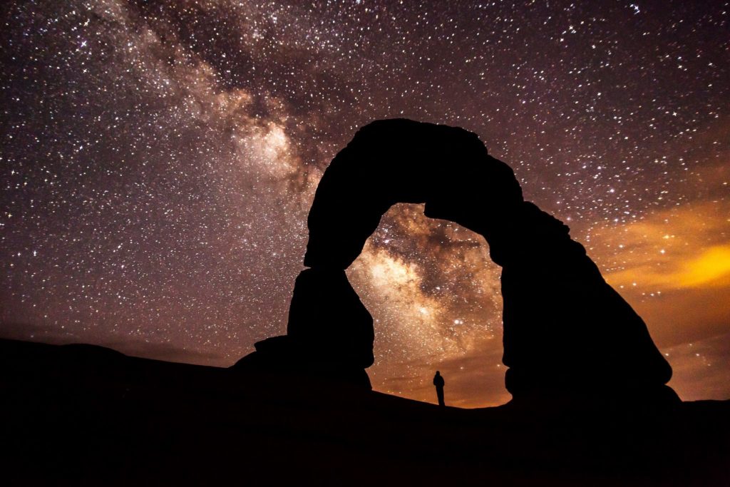 The night sky full of stars with the silhouette of an arch with a person standing underneath.