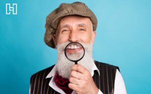 Senior man with white beard wearing newsboy hat and holding up magnifying glass to teeth